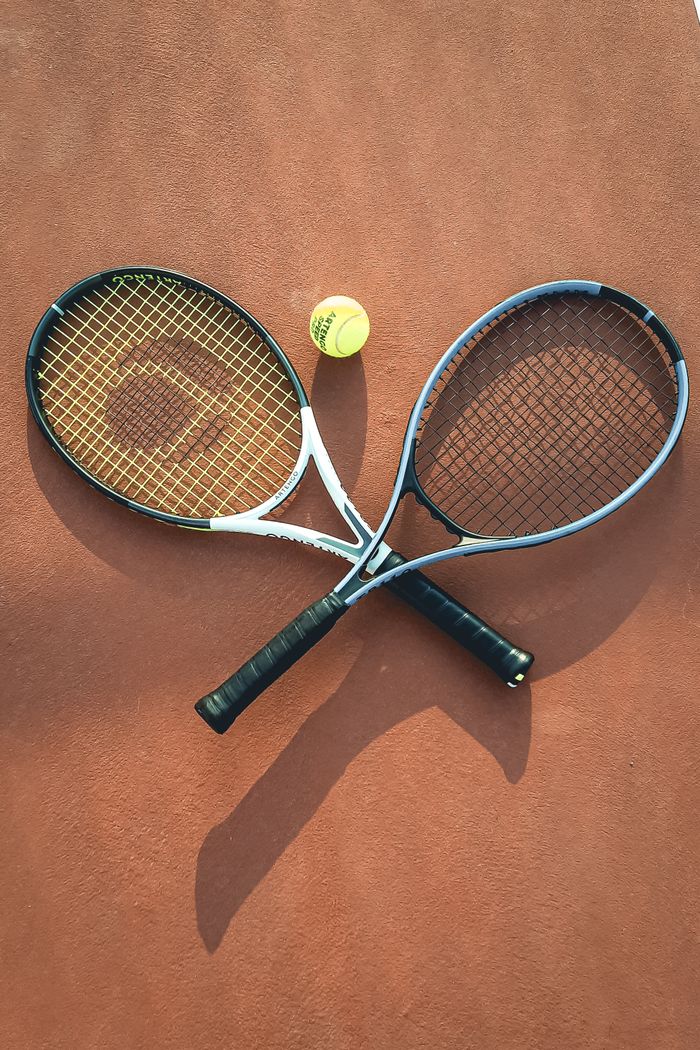 services-03 Two tennis rackets and a ball arranged on a clay court, casting shadows.